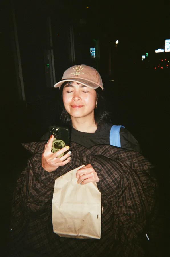 Woman in a baseball cap holds a phone and paper bag on a dark city street at night.