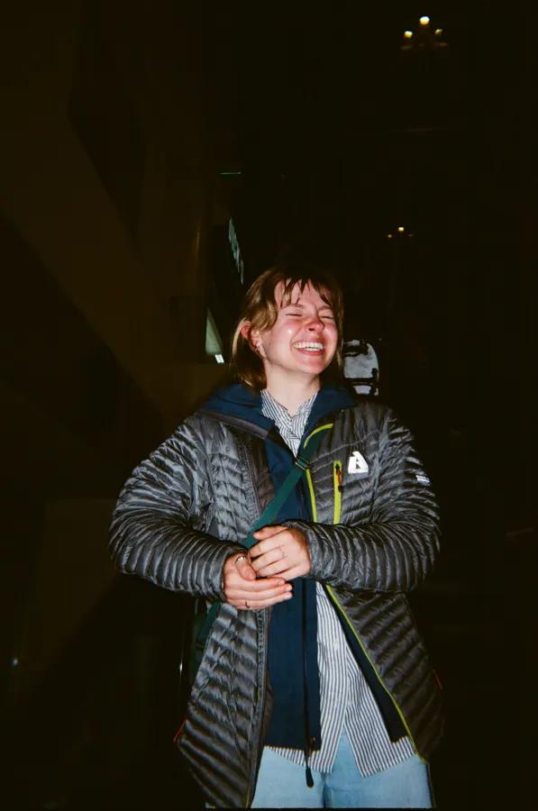 Laughing person in a puffer jacket and scarf stands in a dark room.