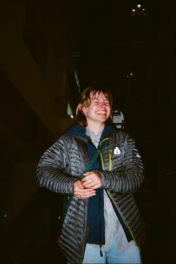 Laughing person in a puffer jacket and scarf stands in a dark room.