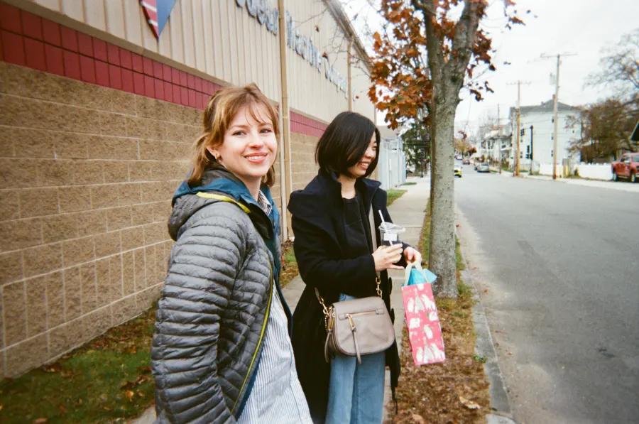 Two women stand on a city sidewalk, one smiling at the camera and the other holding a drink and gift bag.