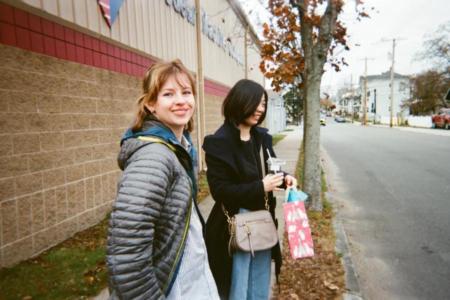 Two women stand on a city sidewalk, one smiling at the camera and the other holding a drink and gift bag.