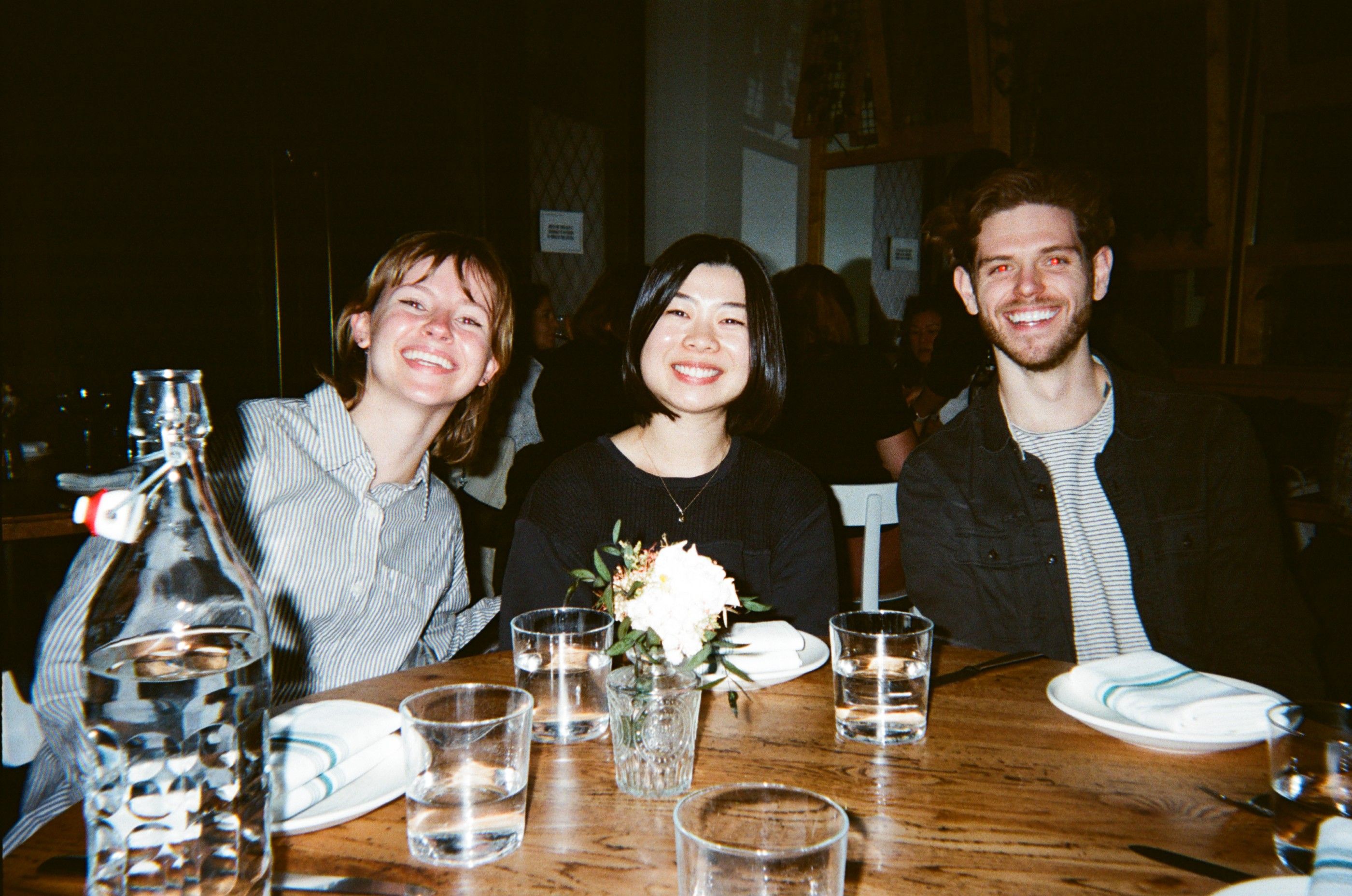 Three smiling friends sit together at a restaurant table set with glasses, plates and flowers.