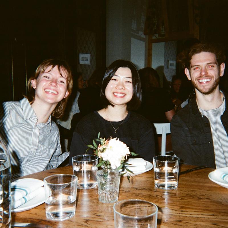 Three smiling friends sit together at a restaurant table set with glasses, plates and flowers.