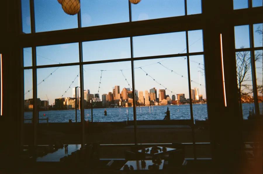 Boston skyline across the harbor, viewed through large restaurant windows at sunset.