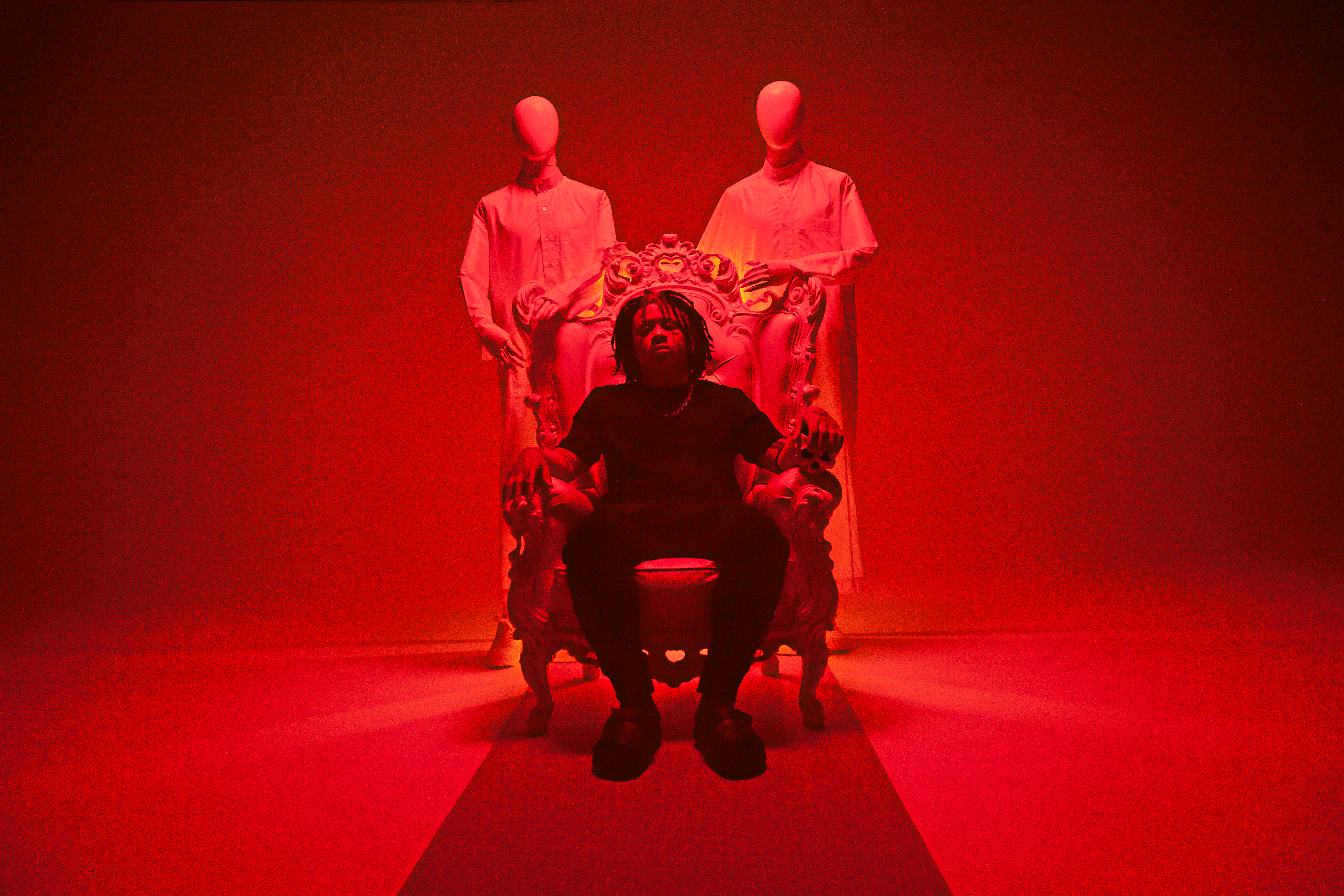 Trippie Redd sits on a production set while bathed in red light glowing from all around him. He has faceless humanoids standing behind him.