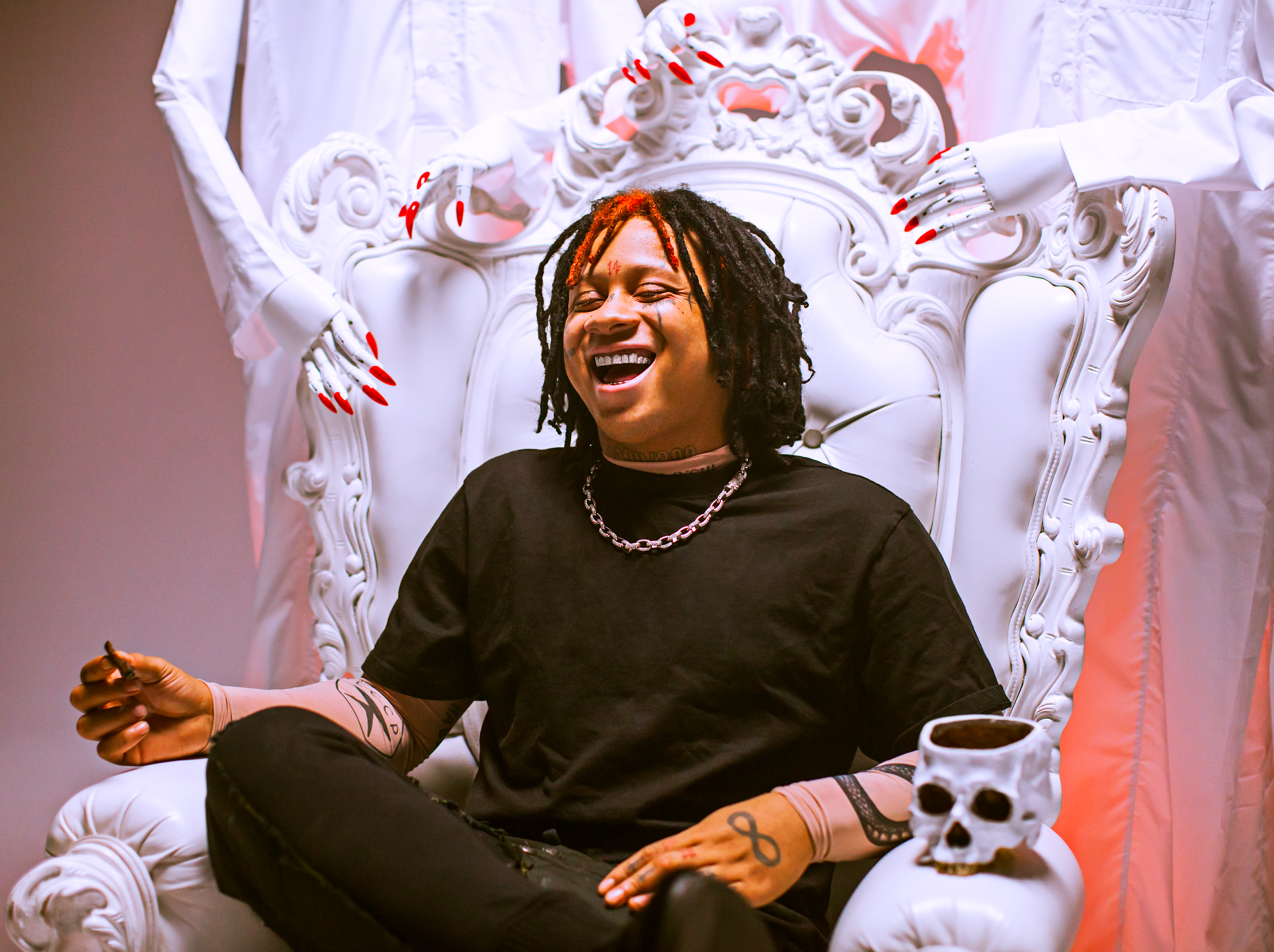Trippie Redd laughing while sitting in chair with long nailed, faceless humanoids stand behind him