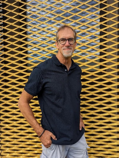 A smiling man with glasses, hands in pockets and a grey beard.