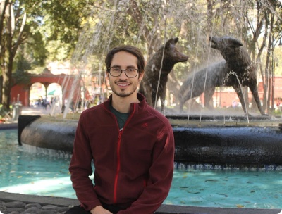 A man with glasses smiling next to a fountain on a sunny day.