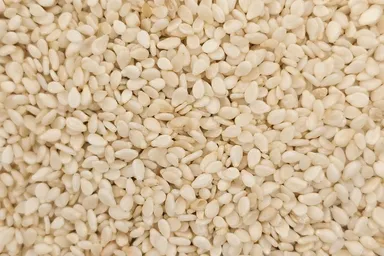 Close-up view of a large pile of hulled sesame seeds, small and oval-shaped, off-white in color, densely packed together.