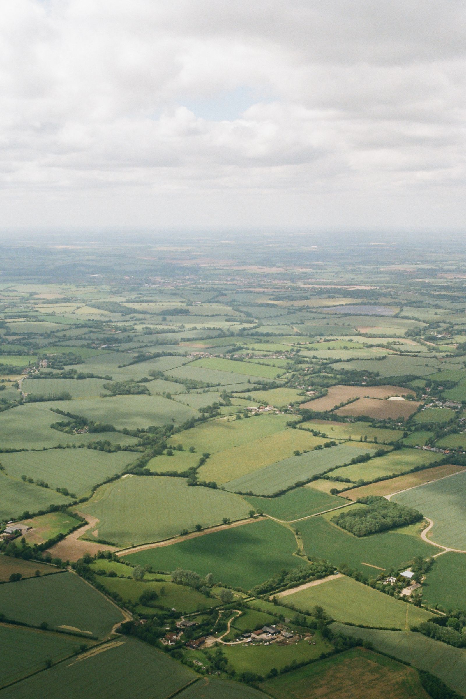 Aerial view of a patchwork of green and brown farm fields divided by hedgerows under a hazy, cloud-filled sky.