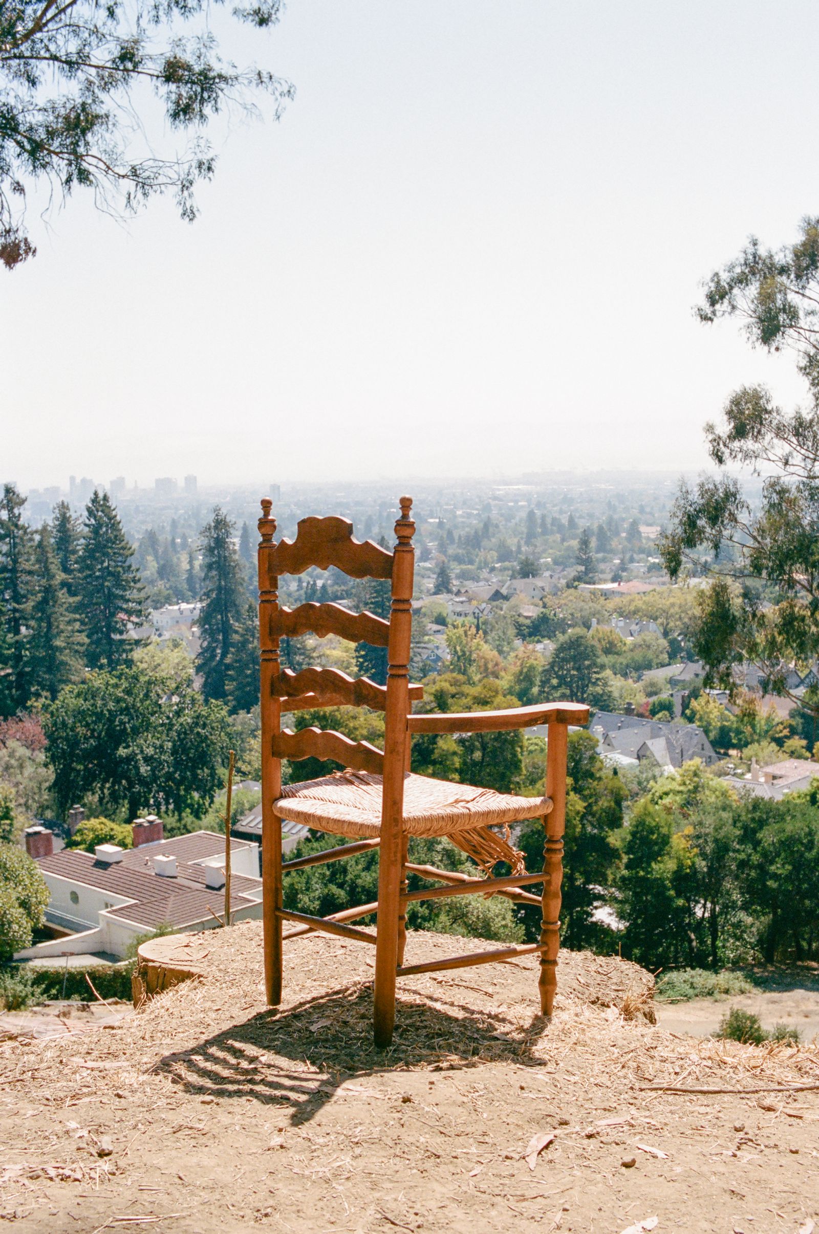 A wooden ladderback chair with a woven rush seat stands alone on a bare dirt clearing above a hazy city and bay.