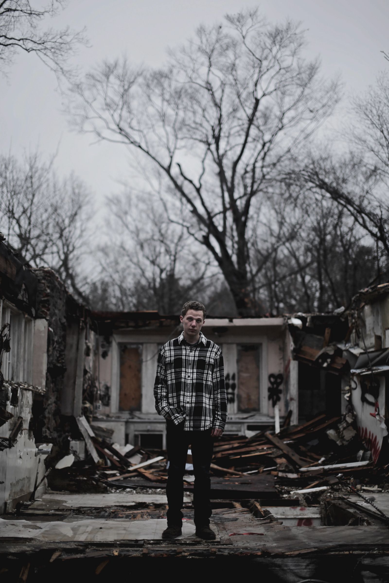 A young man in a plaid flannel shirt stands amid the burned, collapsed ruins of a building with bare trees behind him.