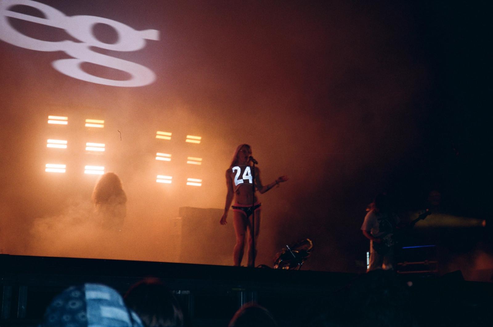 Female performer in a numbered crop top sings into a microphone on a smoky, orange-lit concert stage with bar-shaped lights.
