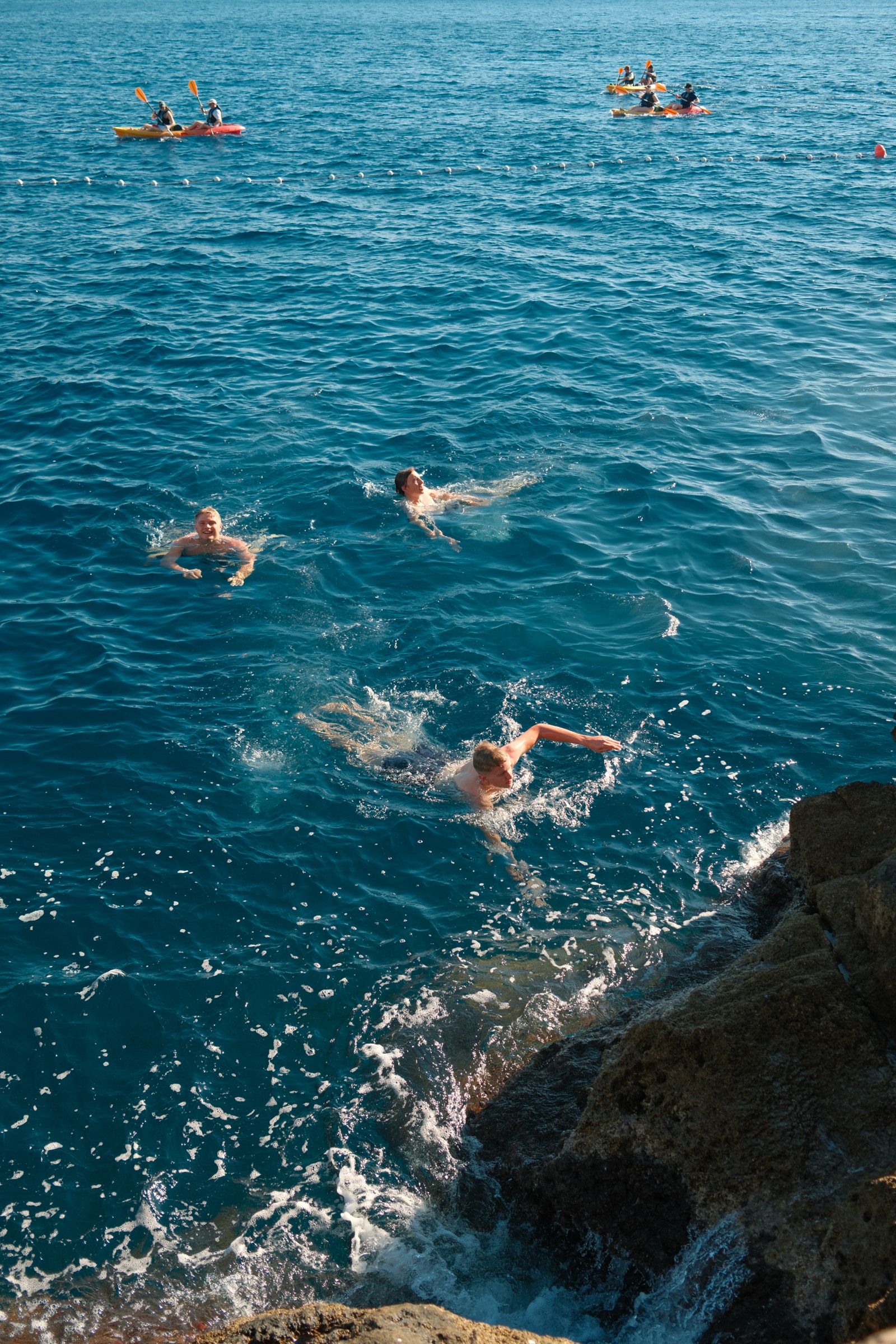 Three swimmers in turquoise sea water near rocks, with two kayaks and paddlers in the background beyond a buoy line.