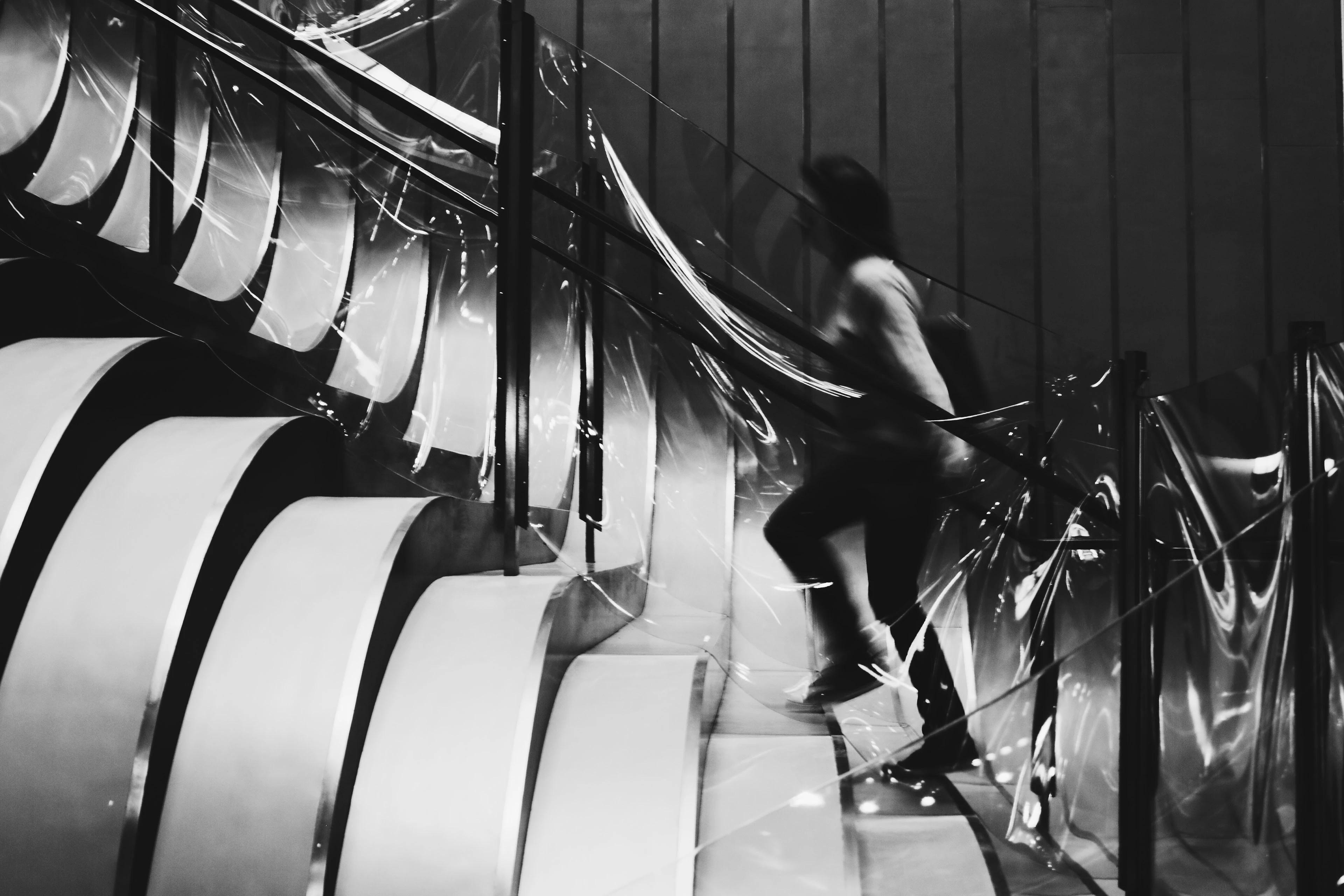 Black-and-white motion-blurred shot of a person ascending a curved staircase between reflective glass panels.