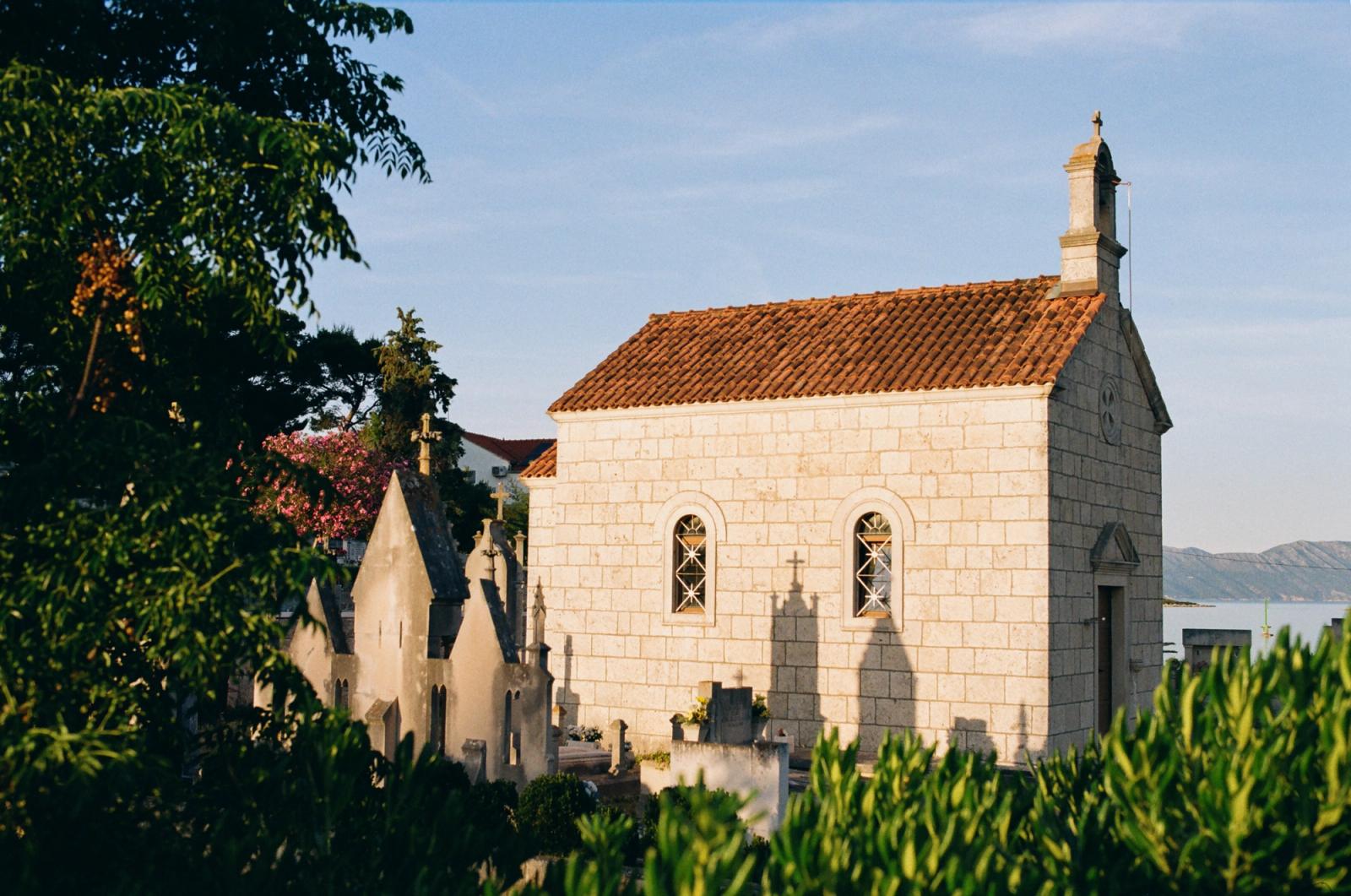 Sunlit stone chapel with a terracotta roof and bell tower stands above a small hillside cemetery of weathered gravestones near the sea.