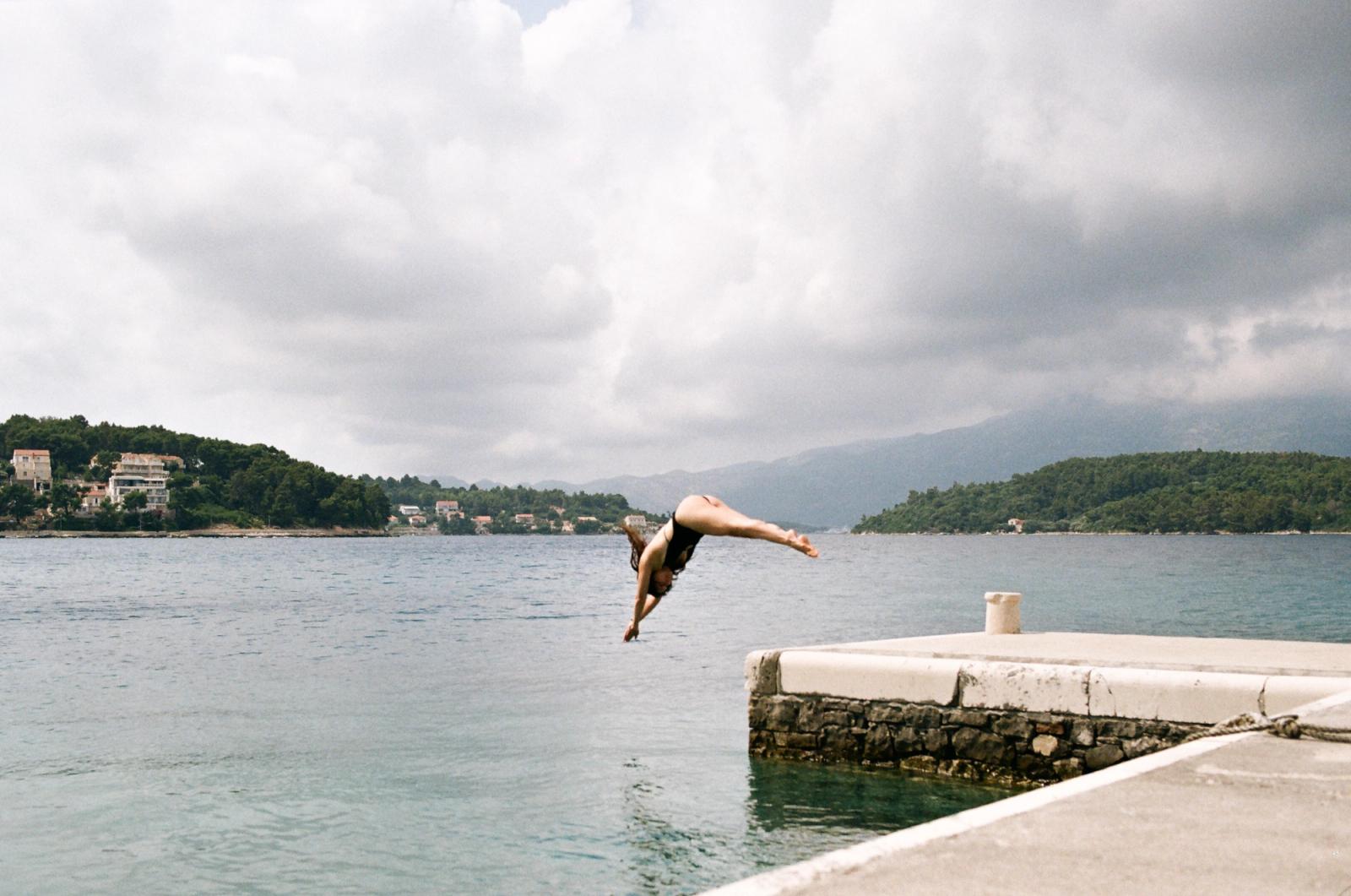 A woman in a black swimsuit dives head-first off a stone pier into calm sea water beneath a cloudy sky and green hills.