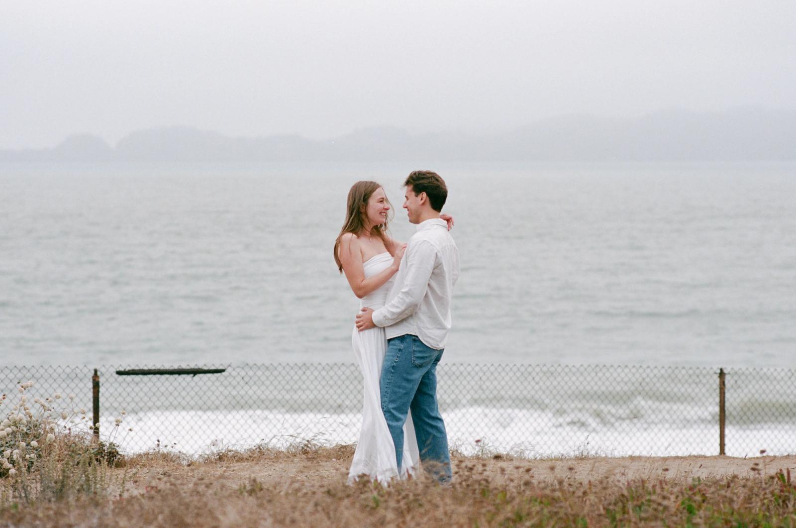 A couple in white stand facing each other in dune grass behind a low chain-link fence, a flat grey bay and hazy headlands stretching out behind them.