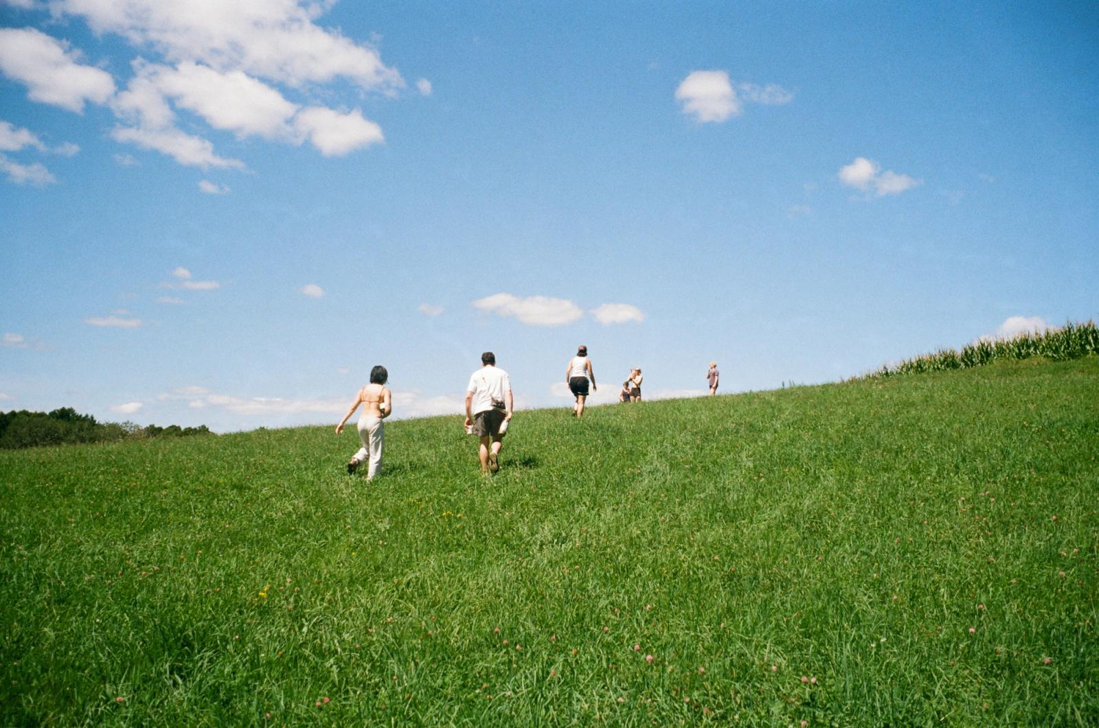 Five people walk away up a steep green hillside toward the crest, small against a bright blue sky streaked with cloud.