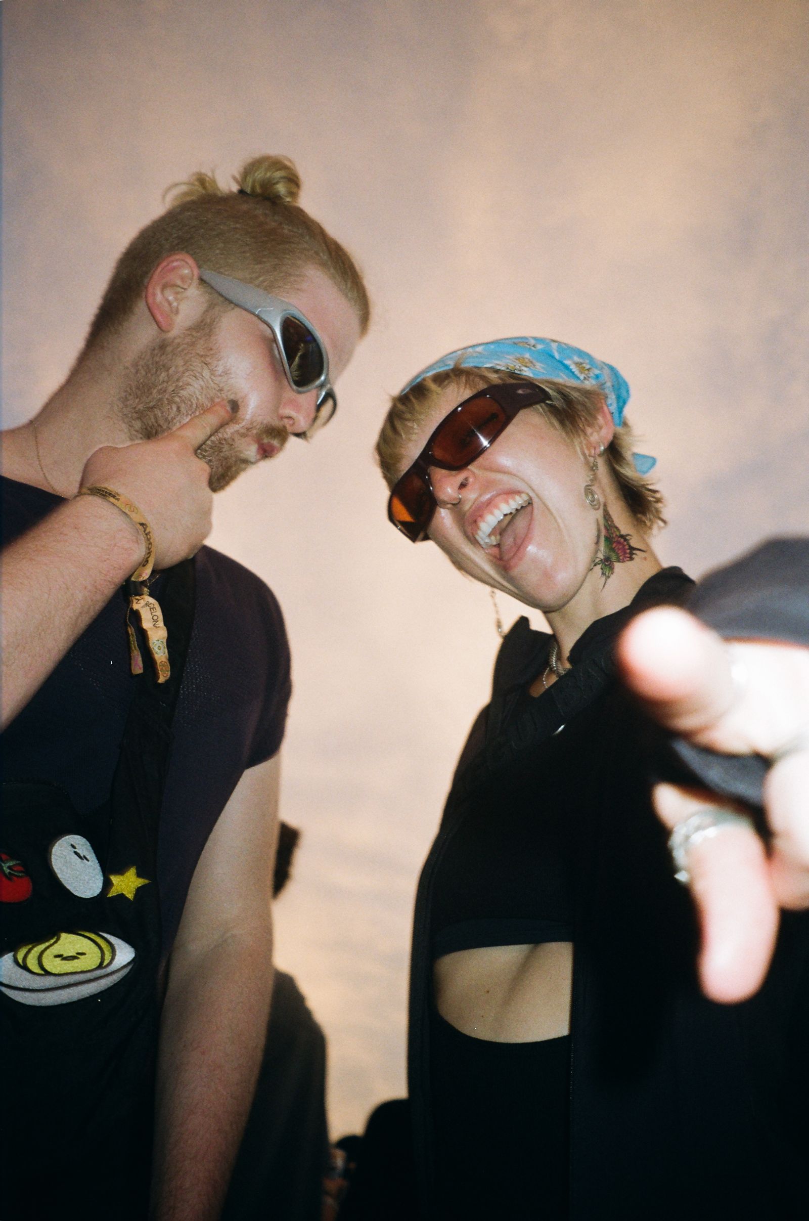 Person in sunglasses points at their cheek beside a laughing woman in sunglasses and a blue bandana, backlit indoors.
