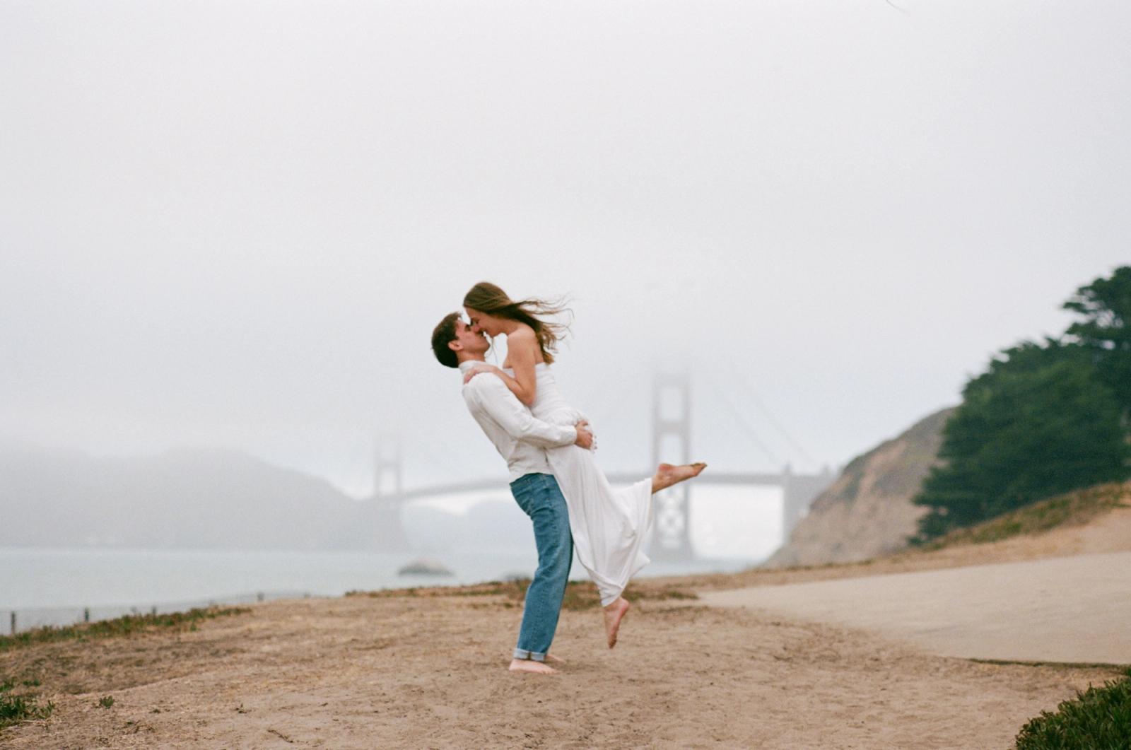 A man lifts a woman off her feet in a kiss on a sandy overlook, her dress and hair swept sideways, the Golden Gate Bridge dissolving into fog behind them.