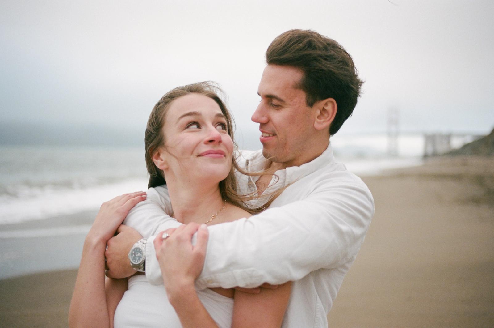 A man in a white shirt wraps his arms around a woman in a white dress on a foggy beach; she tilts her head back to look up at him, both smiling.