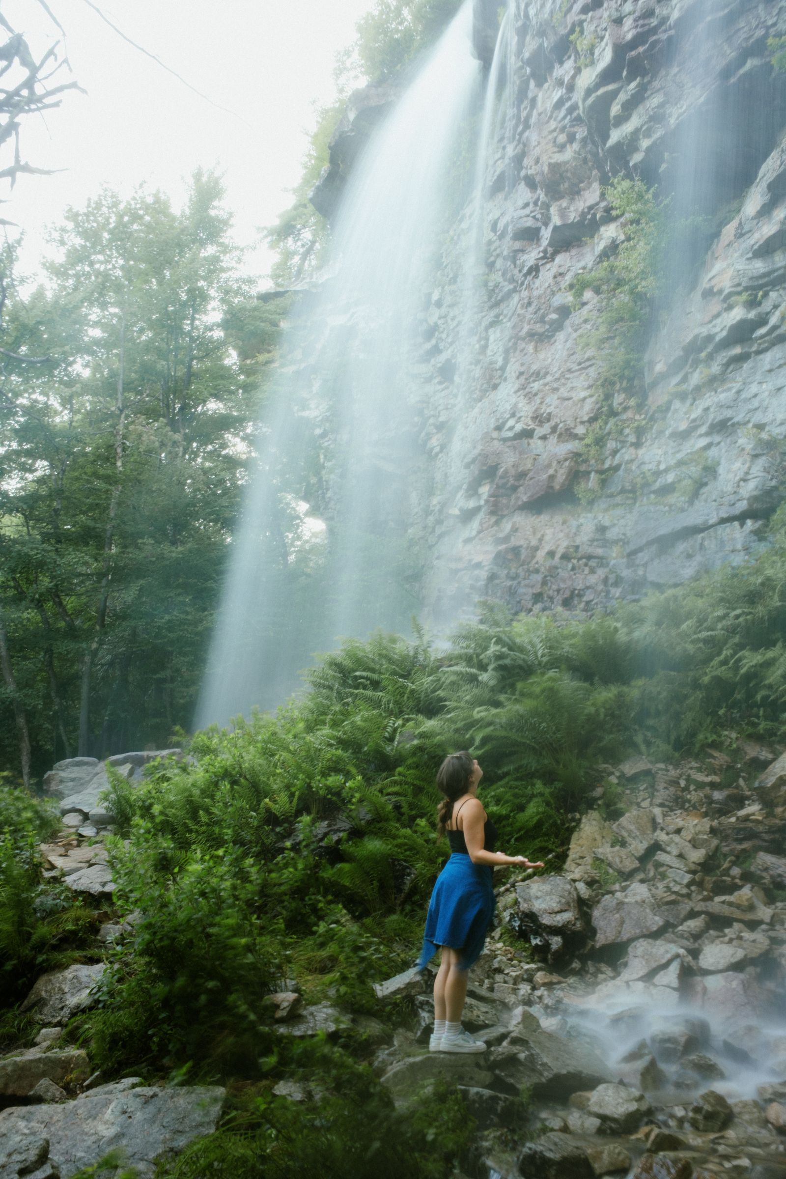 A woman in a black top and blue skirt stands at the base of a waterfall, head tilted back and eyes closed, hands open to the falling mist.