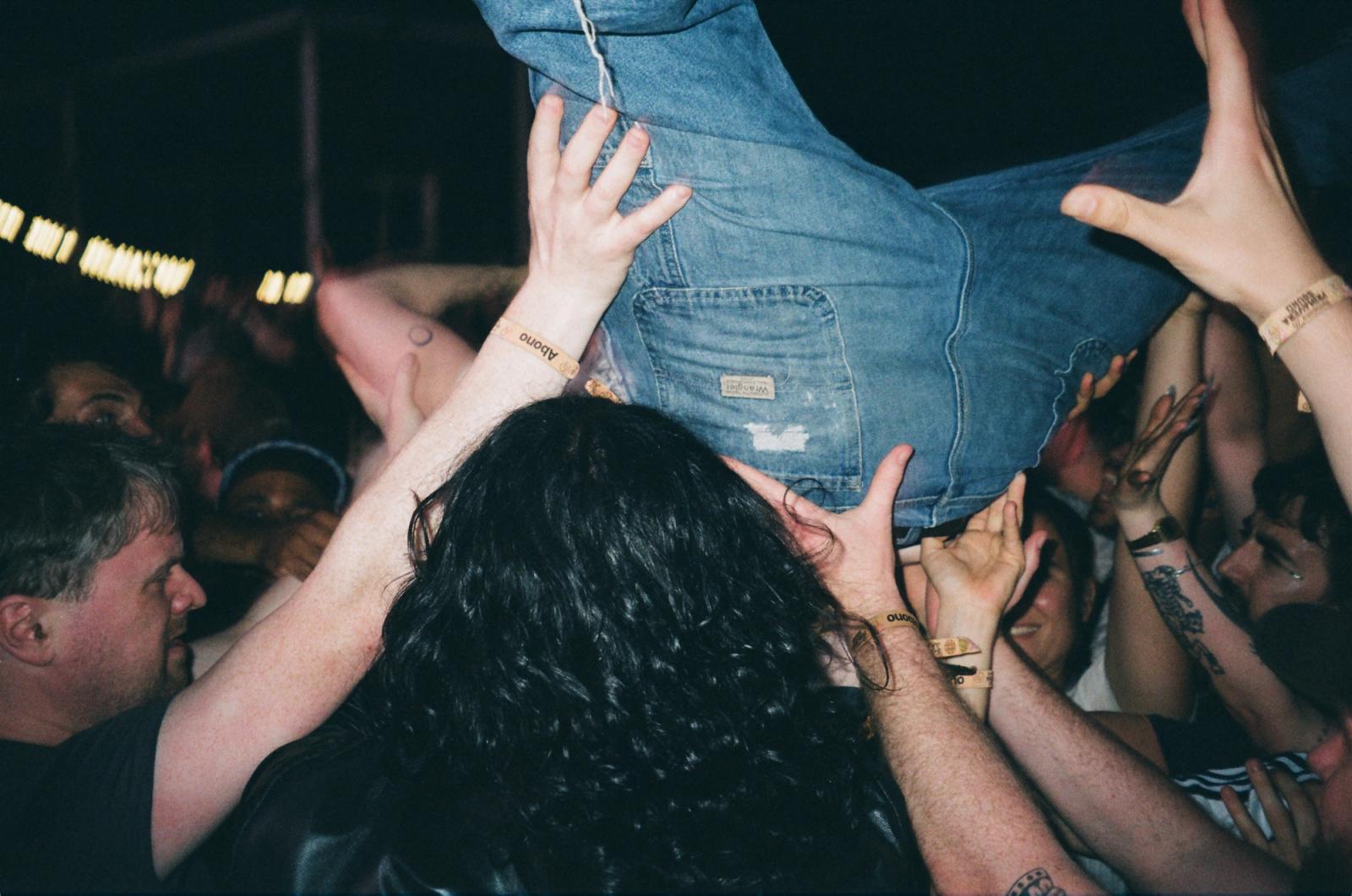 Crowd surfer in blue jeans passes overhead through a sea of raised hands at a packed concert.