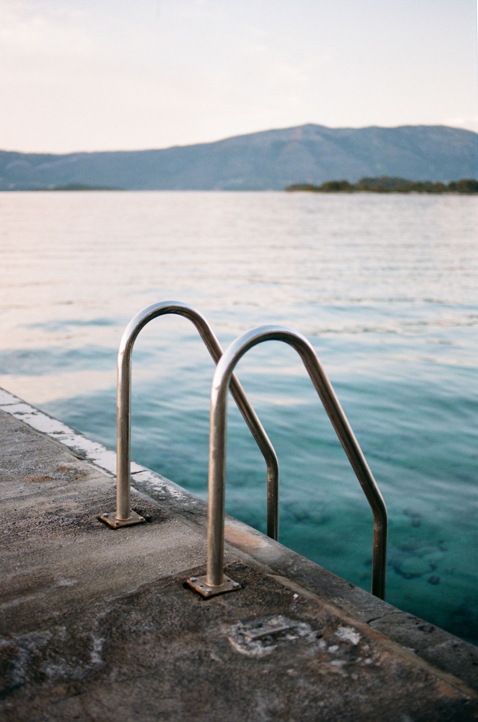 Two stainless steel pool ladder rails curve over the edge of a stone pier into clear turquoise sea, hills in the distance.