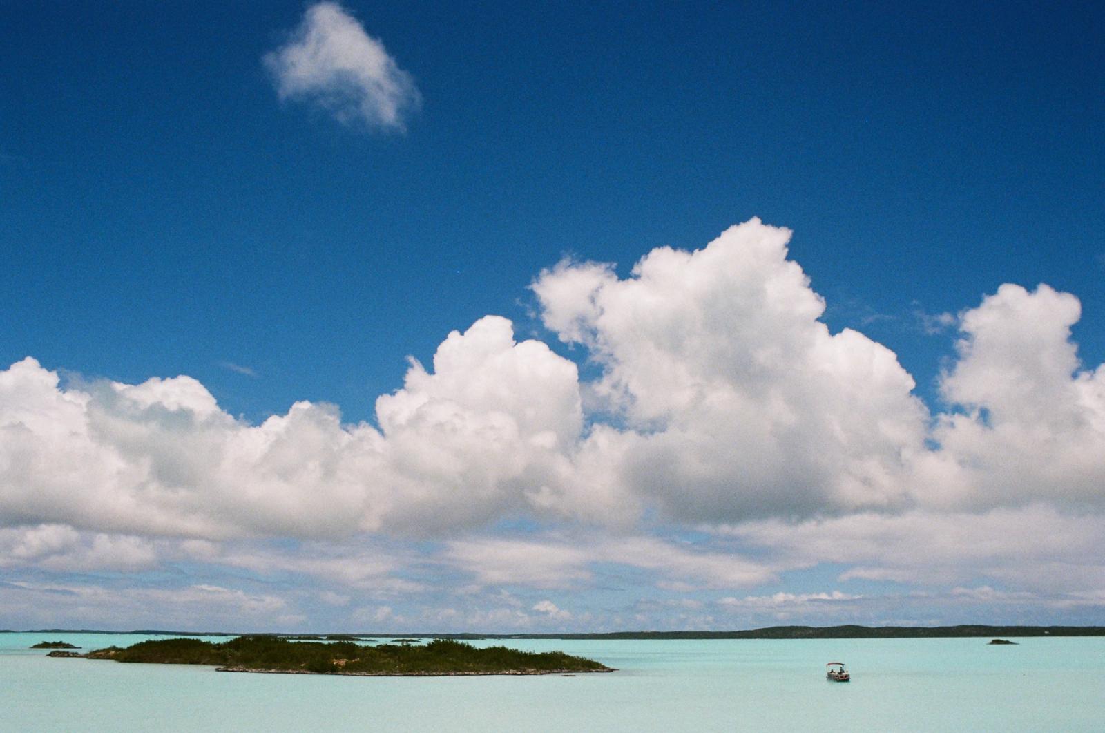 A small boat drifts near low green islands in a bright turquoise lagoon under a blue sky with billowing clouds.