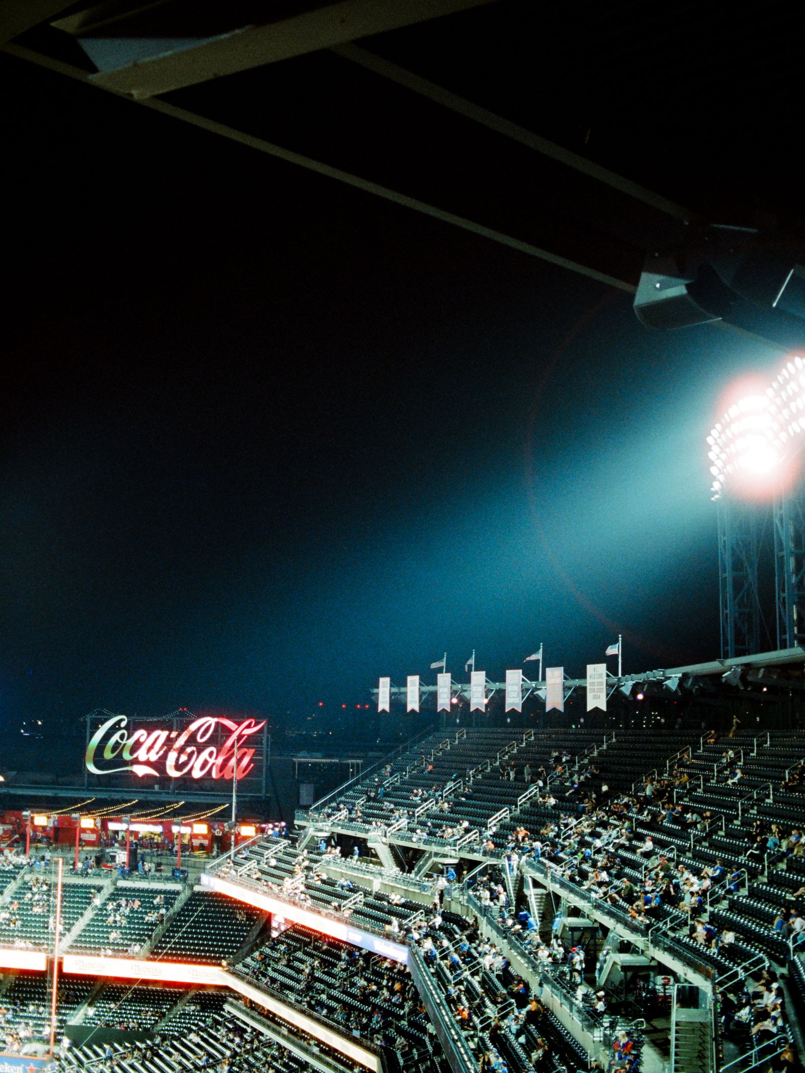 Stadium floodlights flare over the upper deck of a ballpark at night, a neon Coca-Cola sign glowing above the far stands beneath a deep blue-black sky.