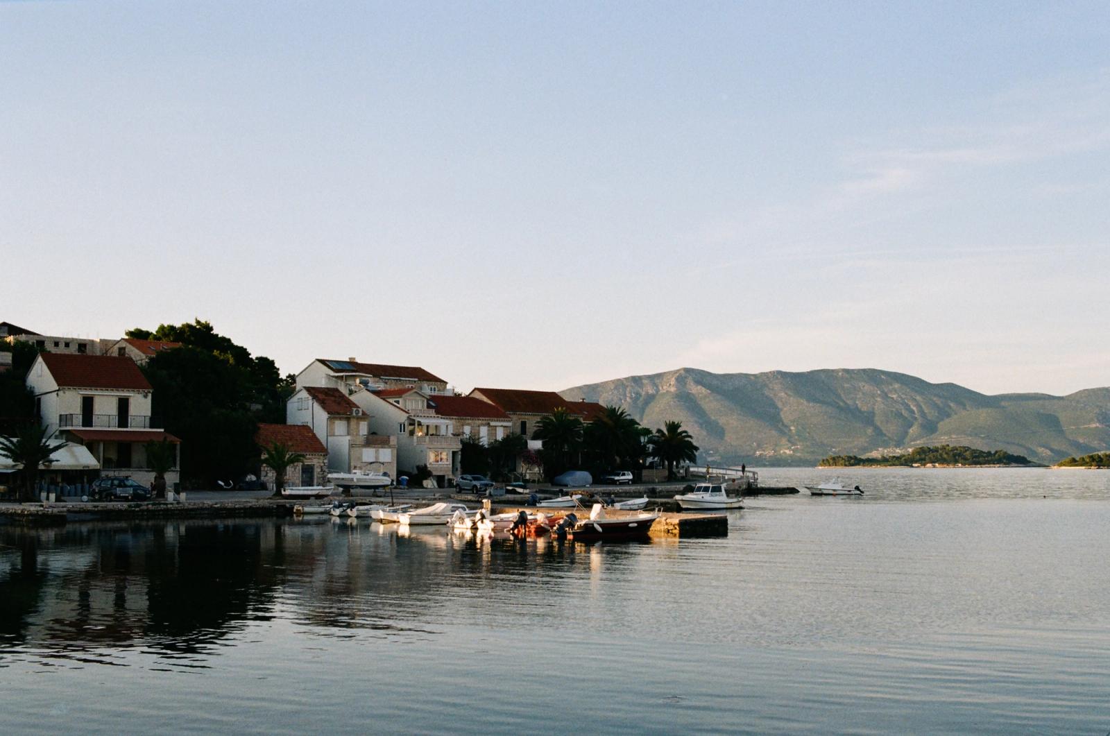 Stone waterfront village with terracotta-roofed houses and moored boats along calm water, mountains rising behind at dusk.