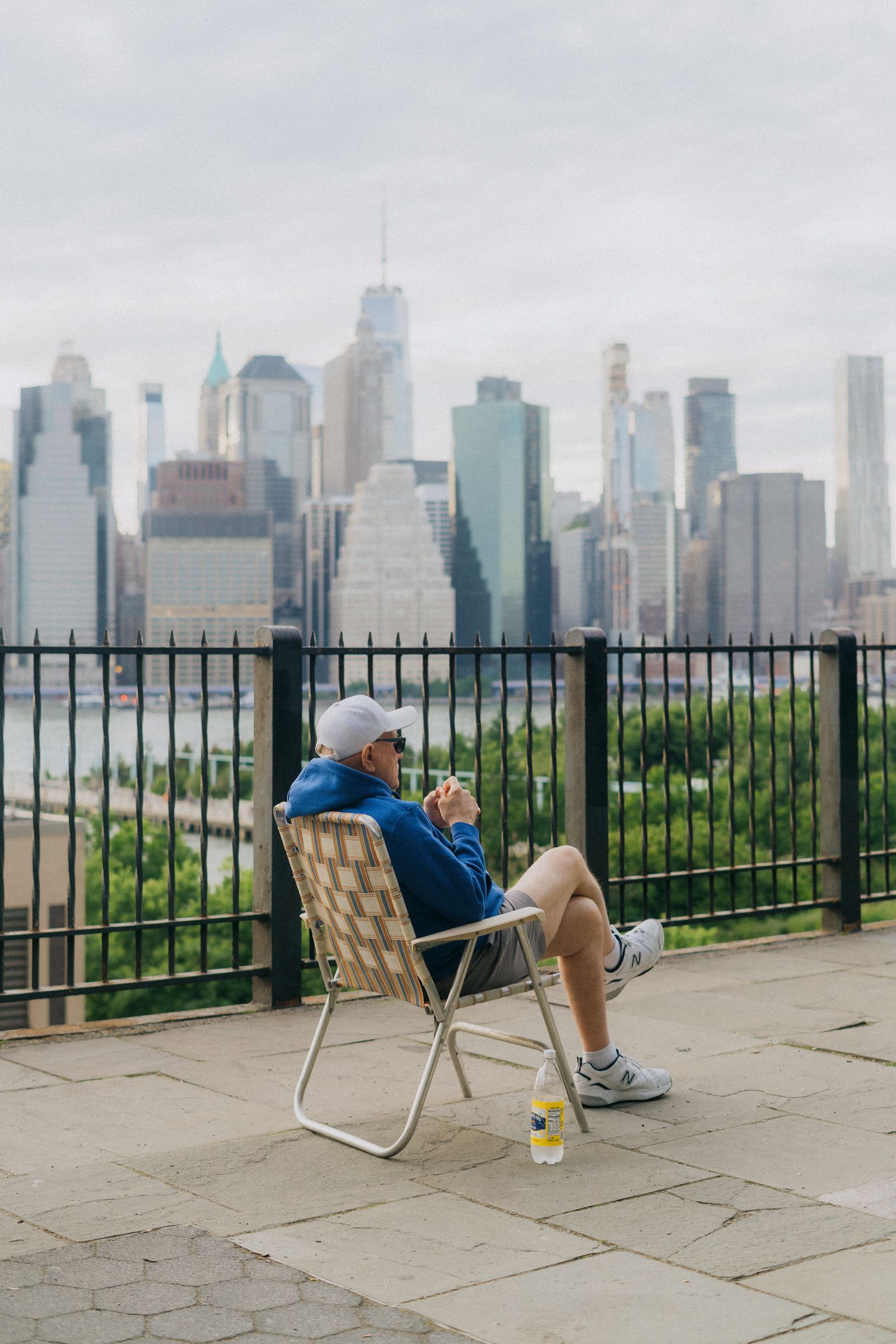 A man in a folding lawn chair looks out over a railing at a dense city skyline across the water.