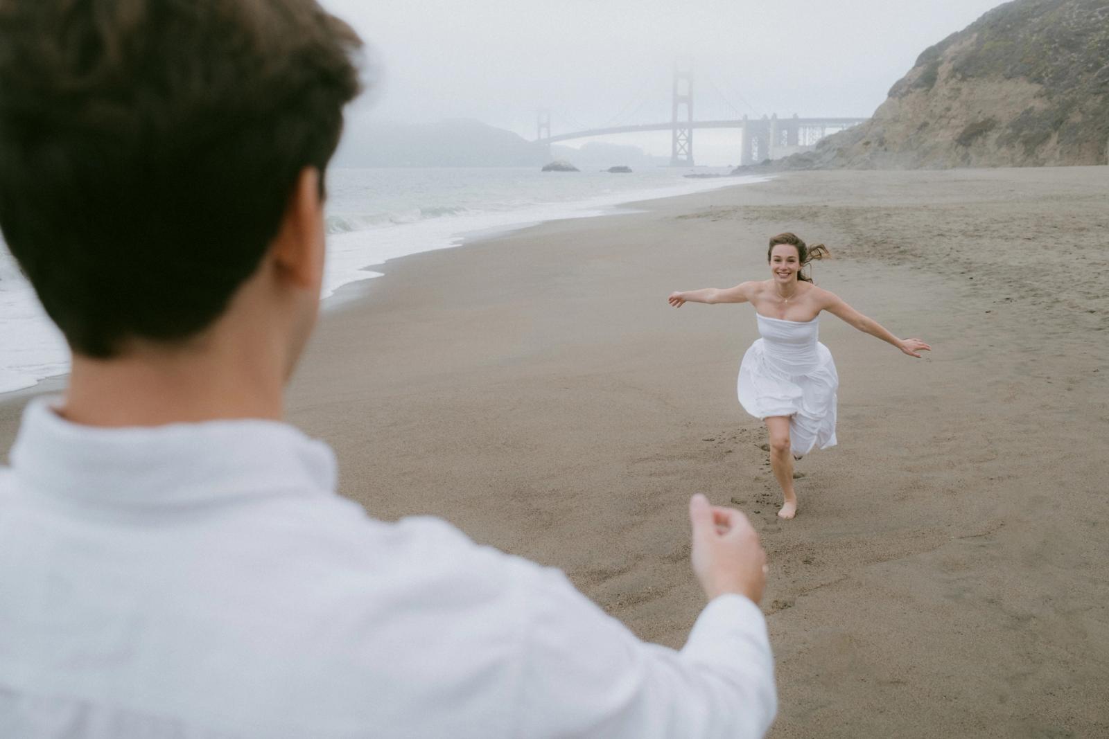 Seen over the shoulder of a man in a white shirt, a woman in a white dress runs barefoot toward him across wet sand, the Golden Gate Bridge faint in the fog behind her.