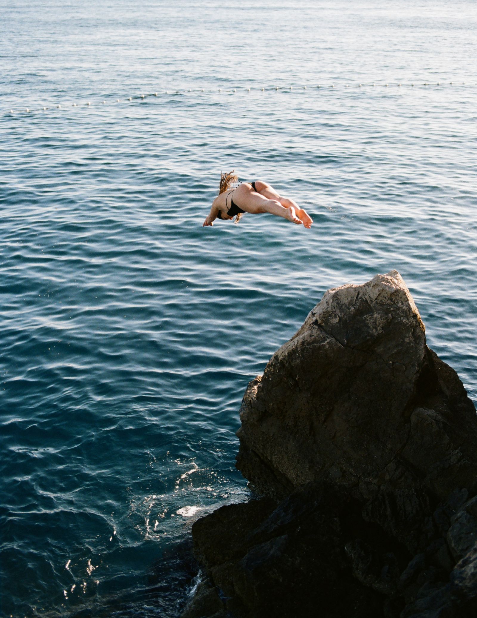 A woman in a black bikini dives head-first through the air toward the sea near a large dark rock, a distant boat on the horizon.