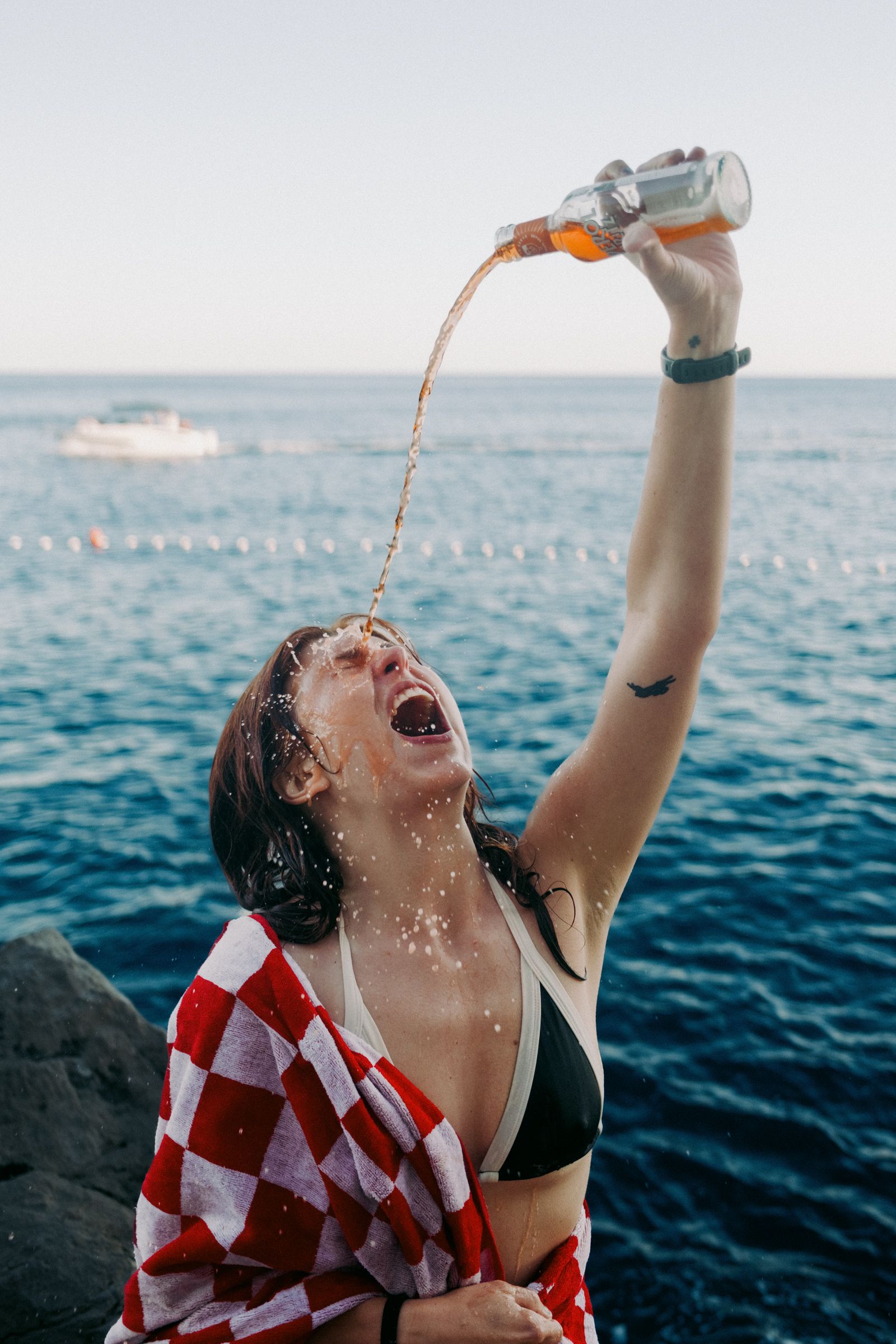 Woman in a bikini wrapped in a red-and-white checked towel pours drink from a bottle into her open mouth, sea behind her.