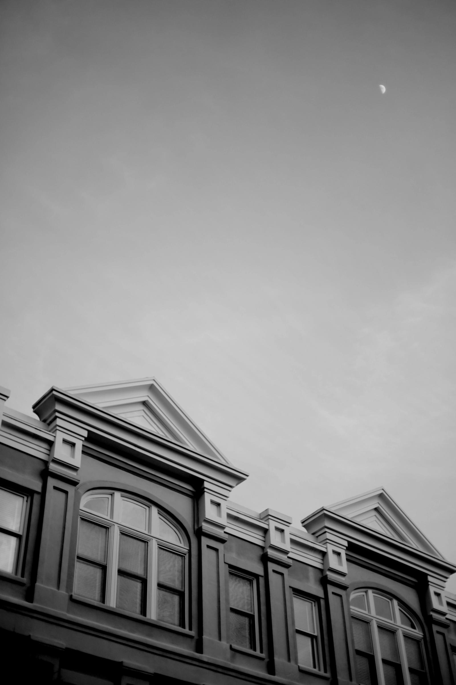 Black-and-white gabled rooftops of a building meet a hazy sky, with a crescent moon visible above.