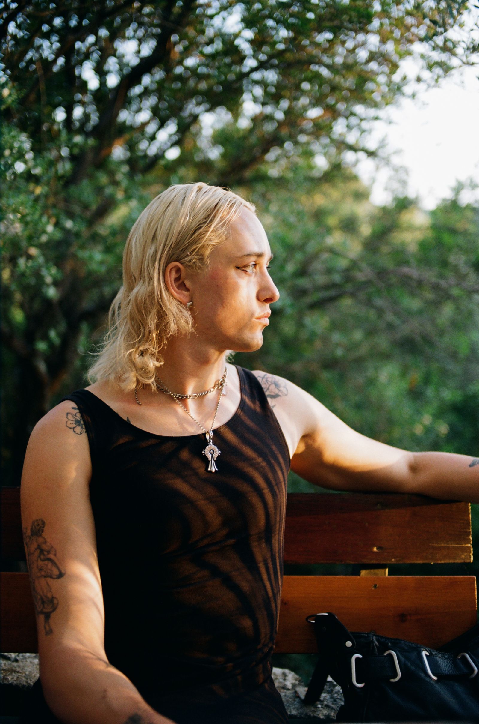 A person with long blond hair and tattooed arms sits on a park bench in golden light, gazing off to the side among trees.