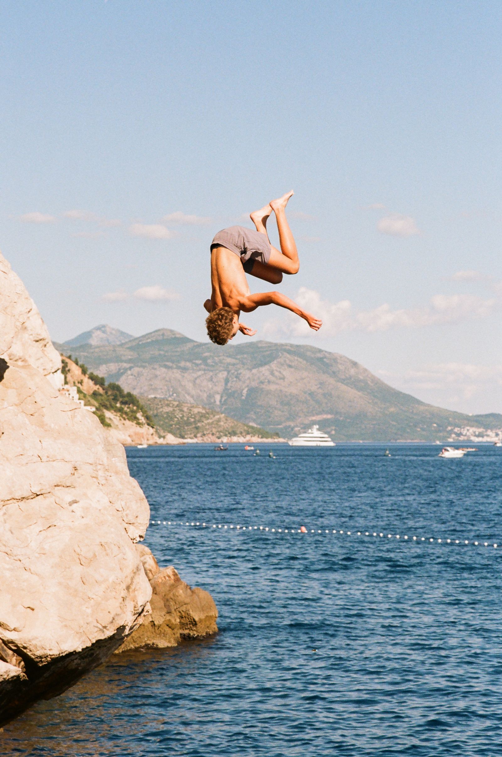 A shirtless man in gray shorts flips backward off a cliff edge, mountains and a yacht visible on the sea behind him.