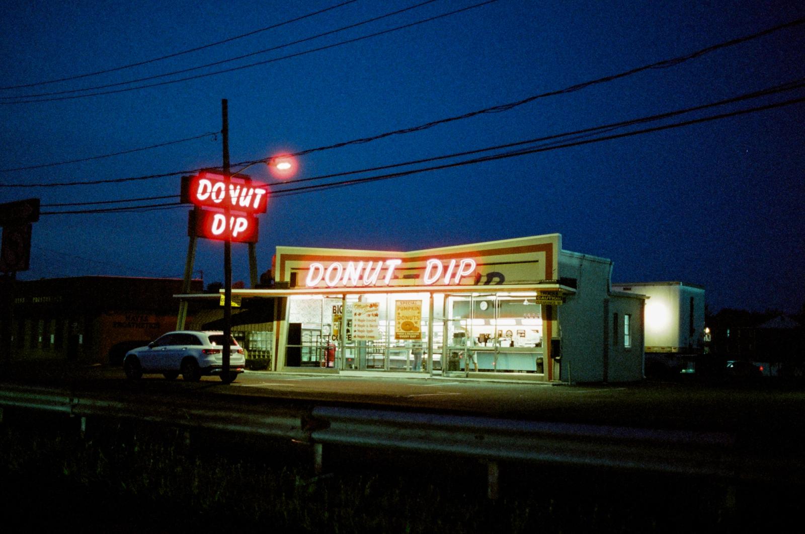 A low roadside donut shop glows red and white against a deep blue dusk sky, its neon sign lit above a lone parked car, power lines crossing overhead.
