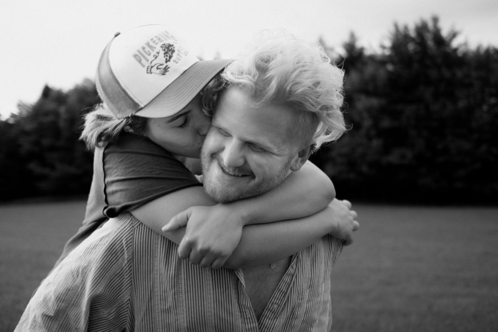 Black-and-white close frame of a woman in a trucker cap with her arms locked around a laughing man’s shoulders, kissing him on the cheek.