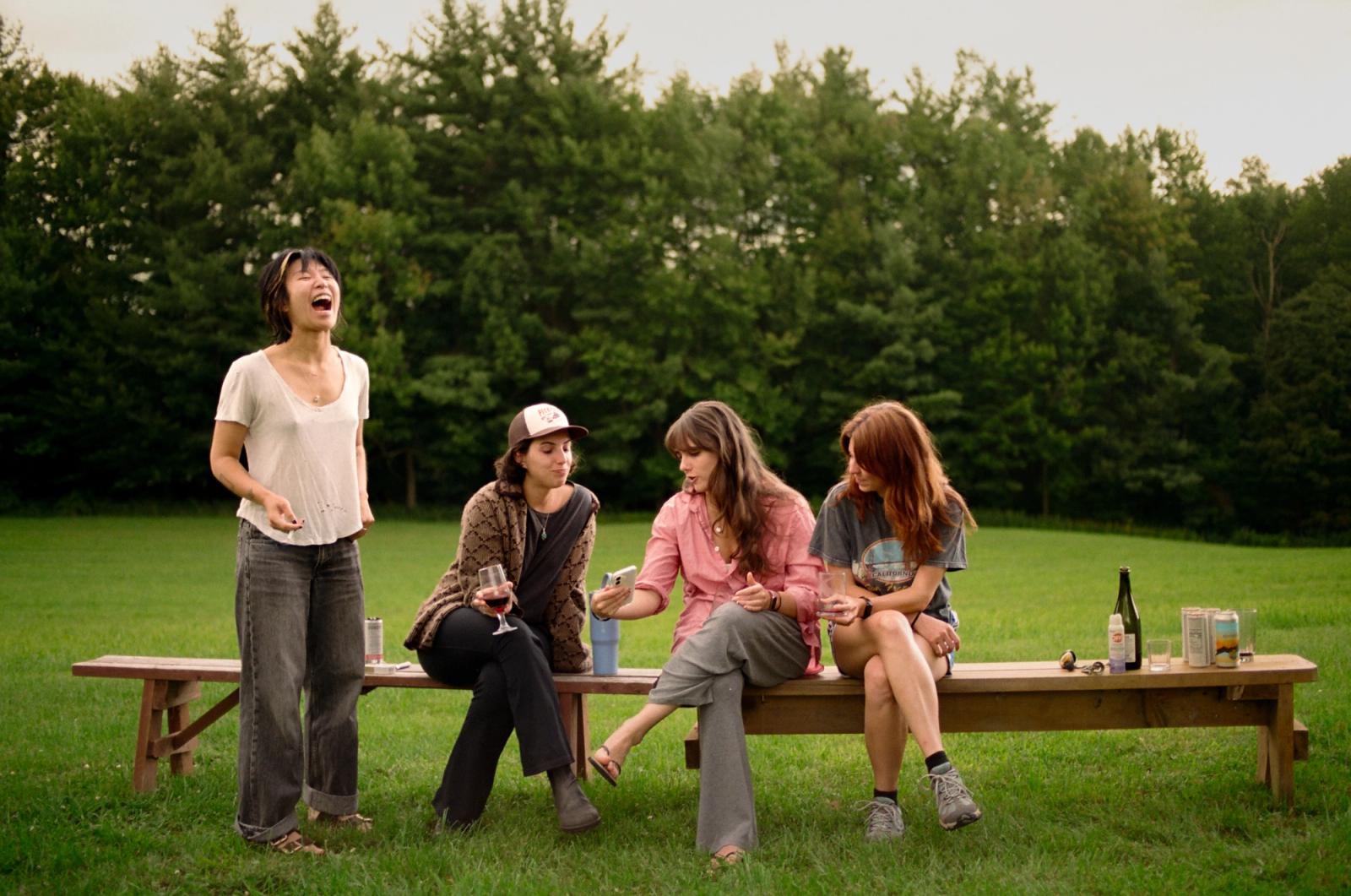Four women sit and stand around a long wooden picnic bench on a lawn at dusk, one laughing with her head thrown back, wine glasses and a bottle on the table.