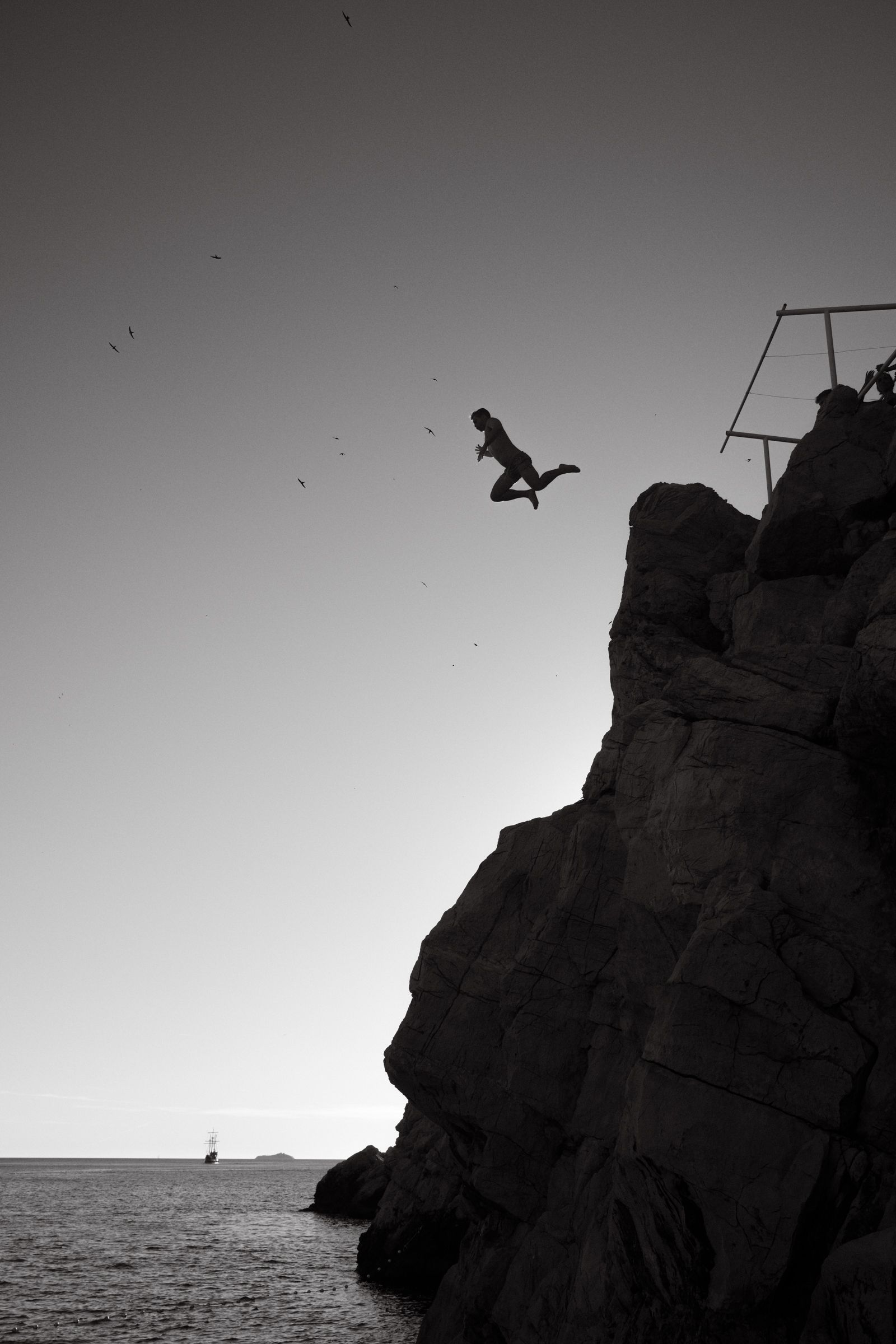 Silhouetted figure leaping from a rocky cliff platform into open air, birds scattered across the sky, a tall ship visible far below.