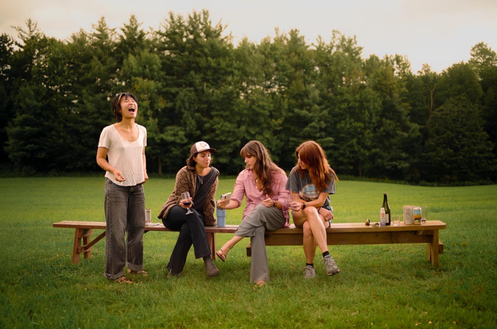 Four women sit and stand around a long wooden picnic bench on a lawn at dusk, one laughing with her head thrown back, wine glasses and a bottle on the table.