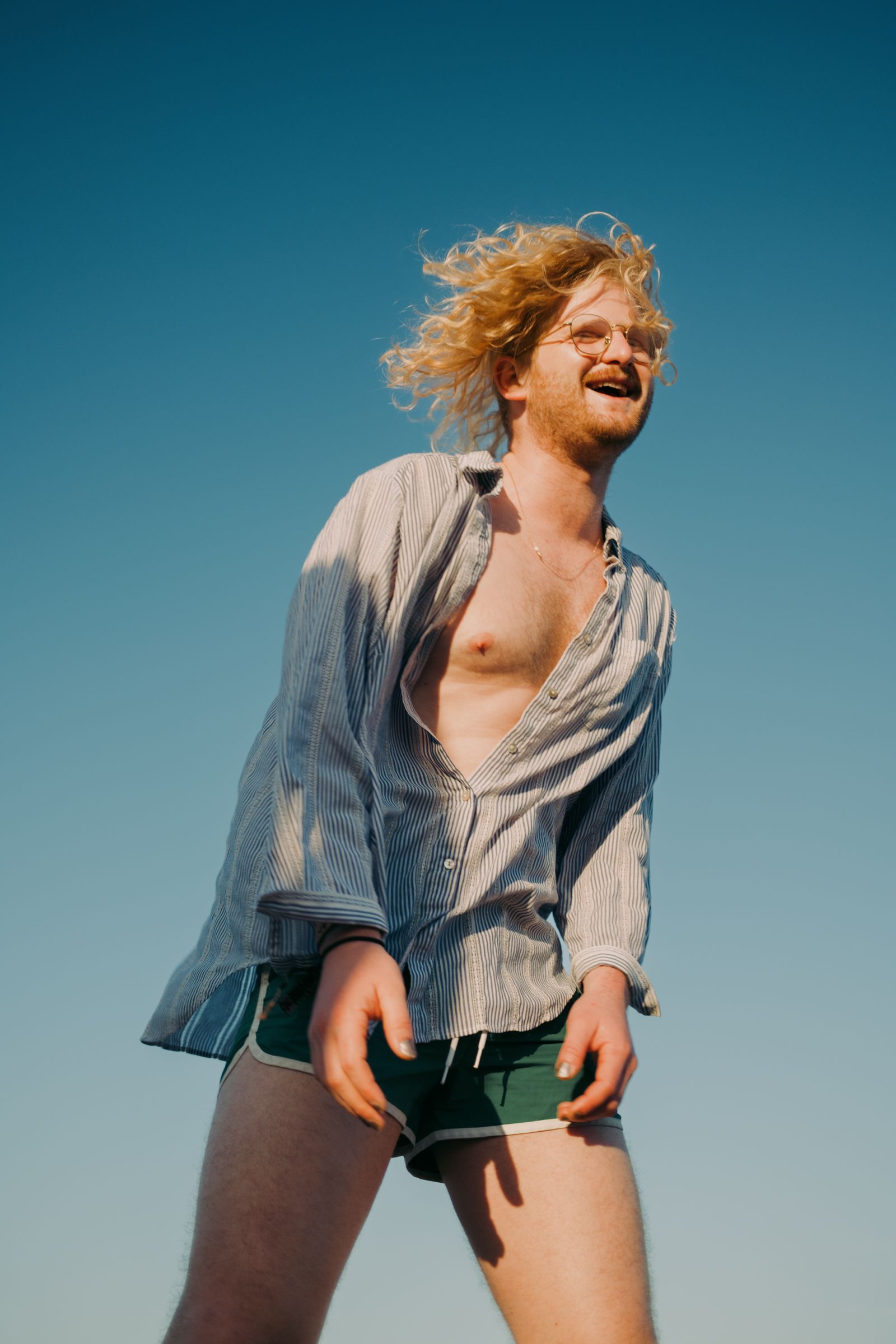 Curly-haired person in an unbuttoned striped shirt laughs against a clear blue sky, wind blowing his hair.