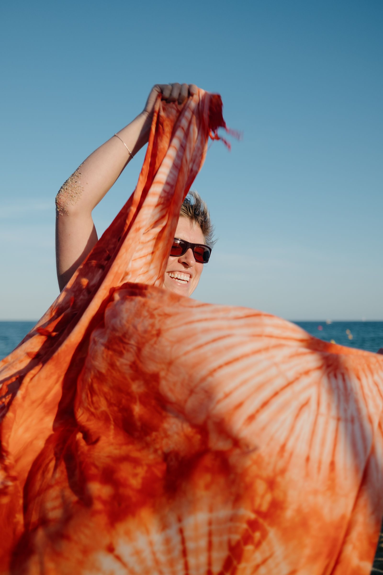 A person in sunglasses holds up a large orange tie-dye fabric overhead, laughing, with the sea and sky behind them.