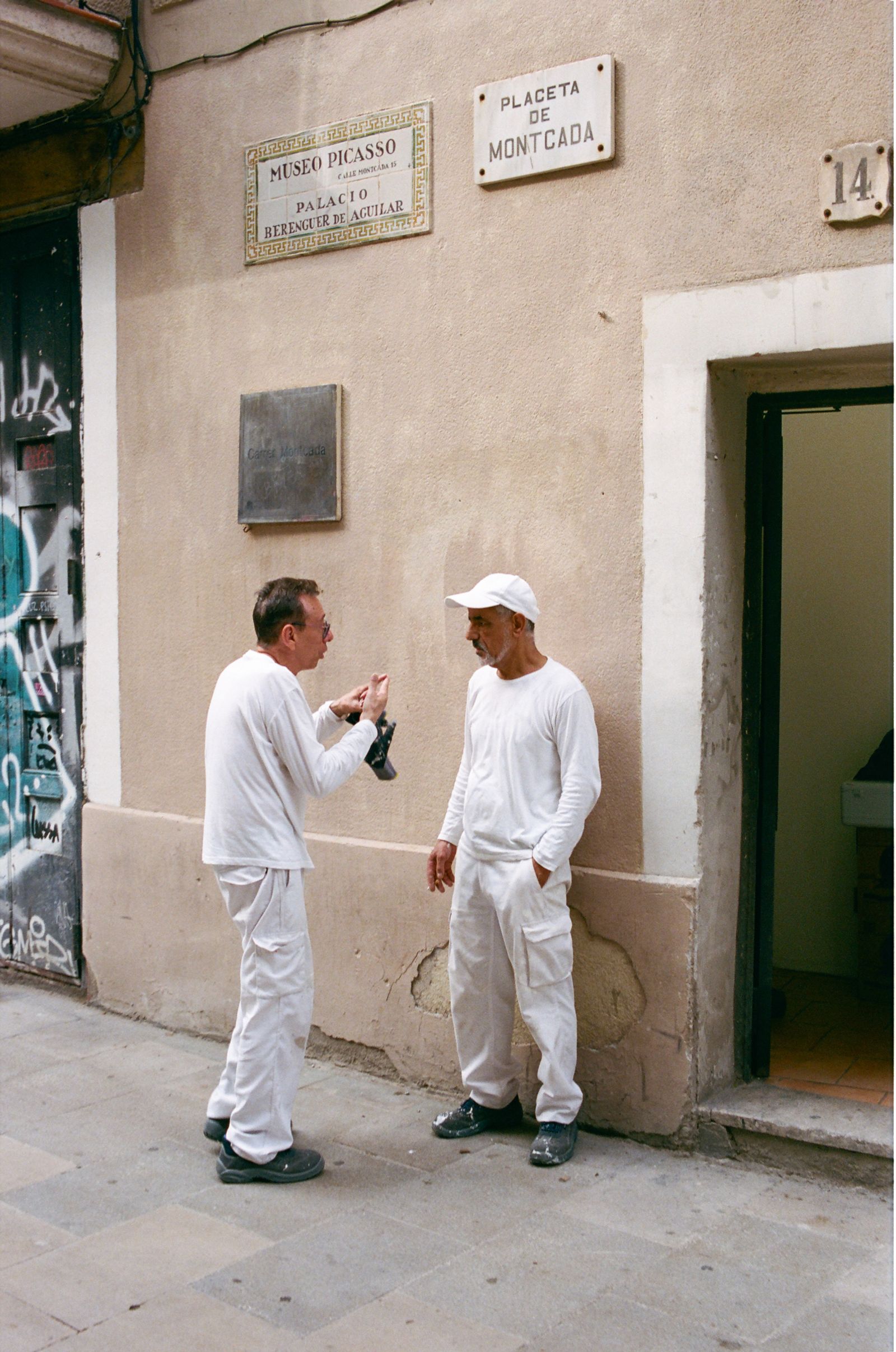 Two men in matching white painter's clothes talk on a stone street beneath signs reading Museo Picasso and Placeta de Montcada.