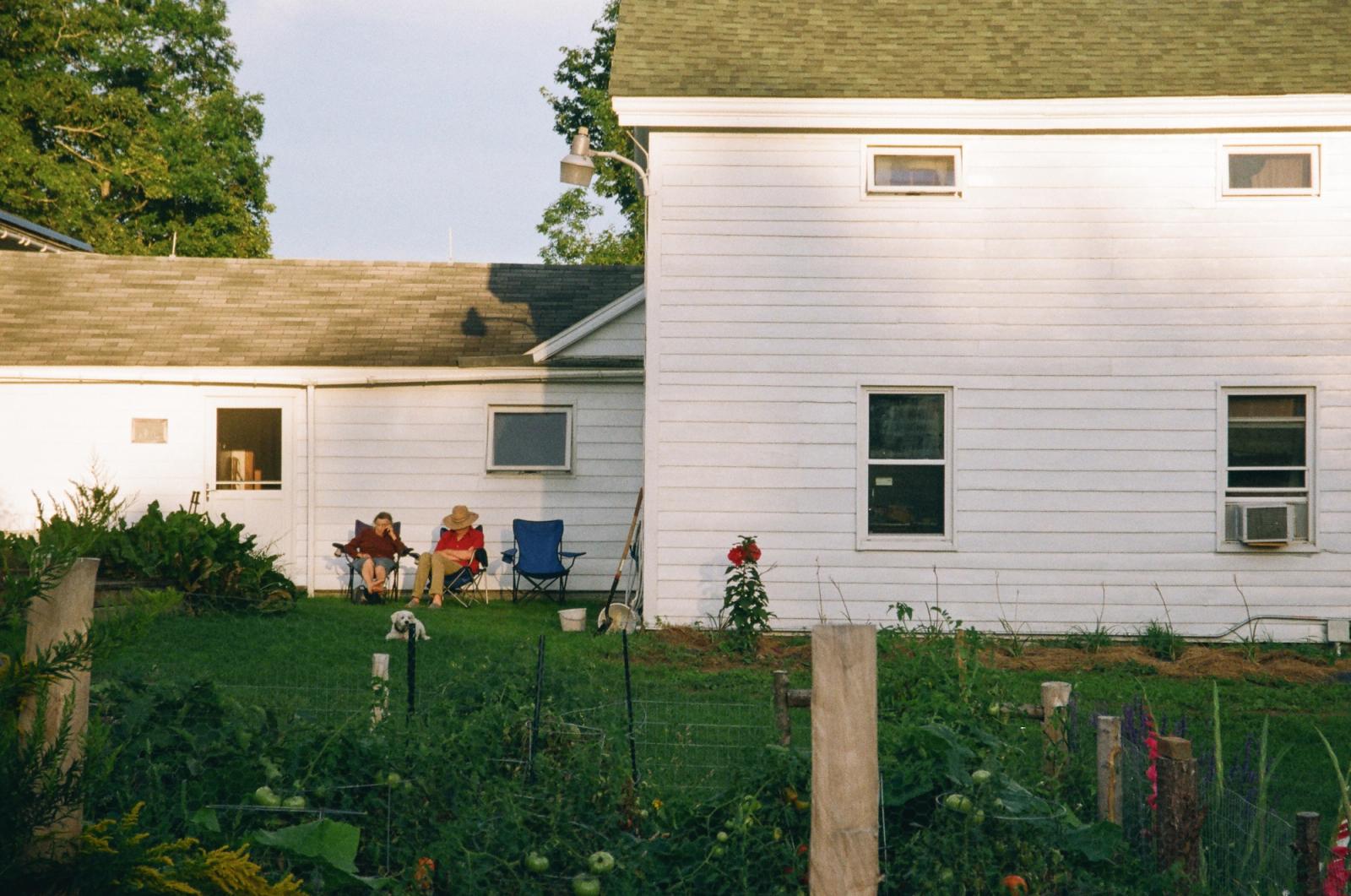 Two people sit in folding lawn chairs in the evening sun beside a white clapboard farmhouse, a fenced vegetable garden heavy with tomatoes in the foreground.