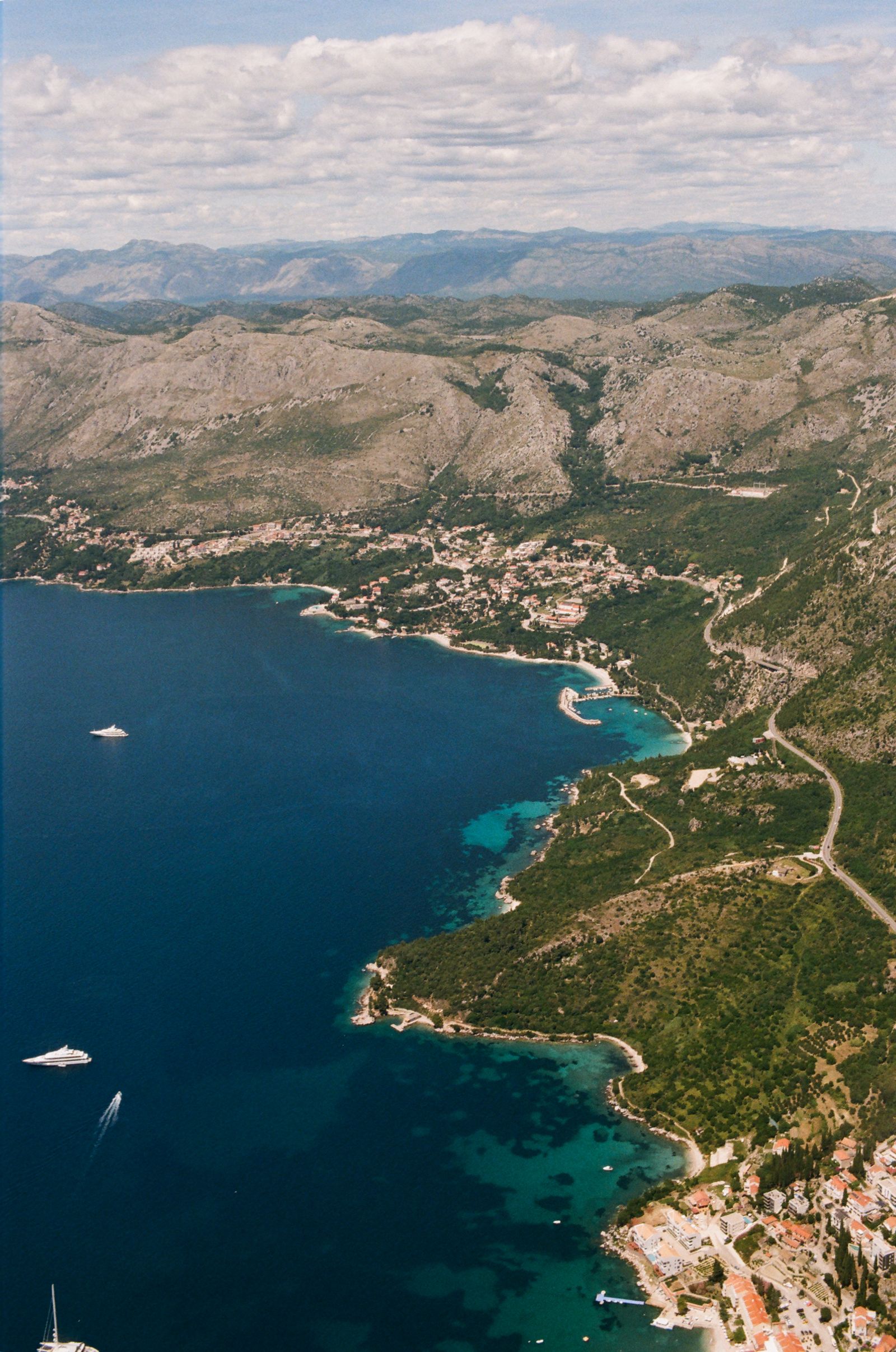 Aerial view of a deep blue bay meeting turquoise shallows along a forested coastline dotted with red-roofed villages and yachts.
