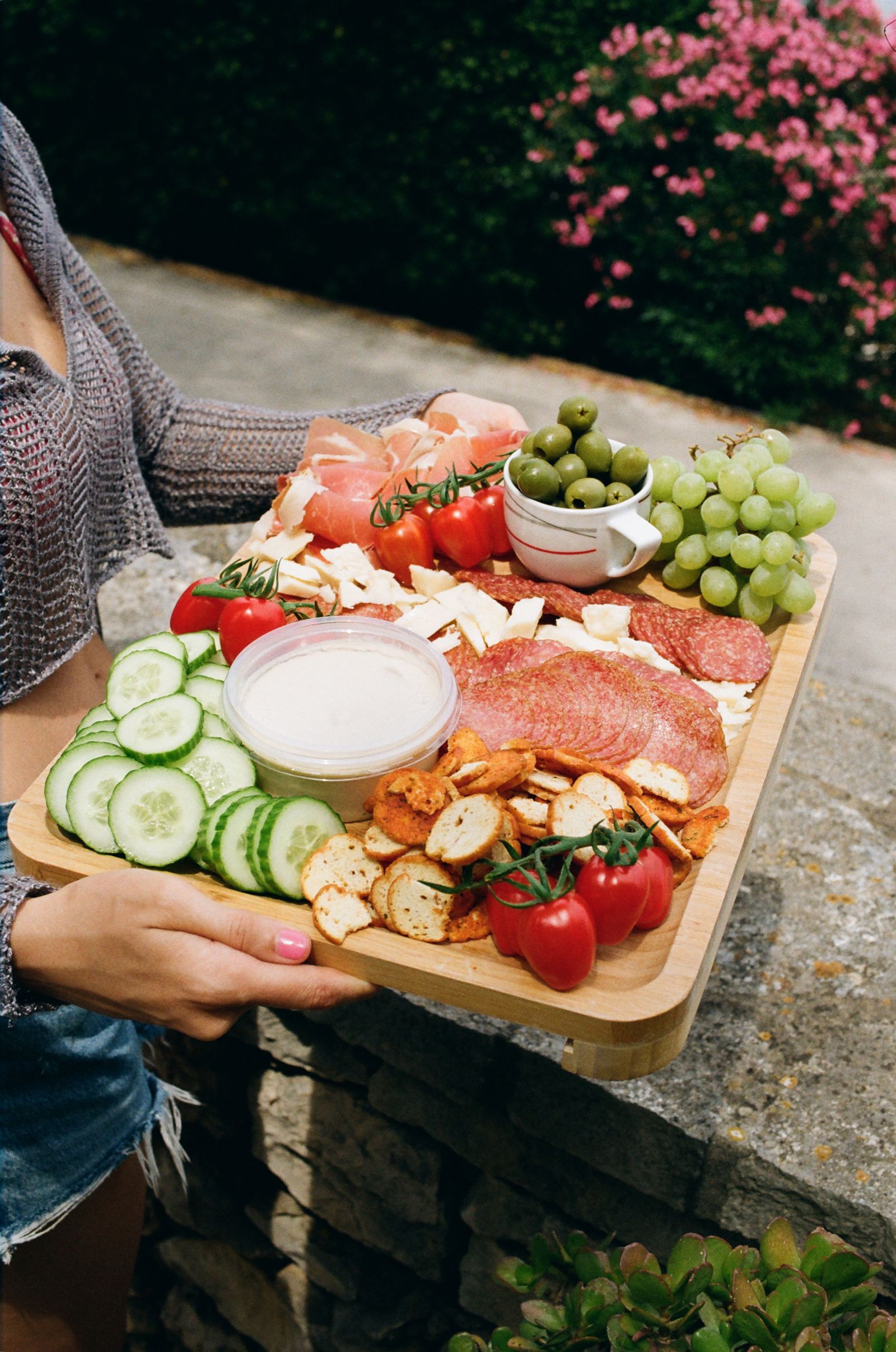 Hands hold a wooden board loaded with cured meats, cheese, olives, grapes, cucumber, tomatoes, crackers, and dip on a stone ledge.